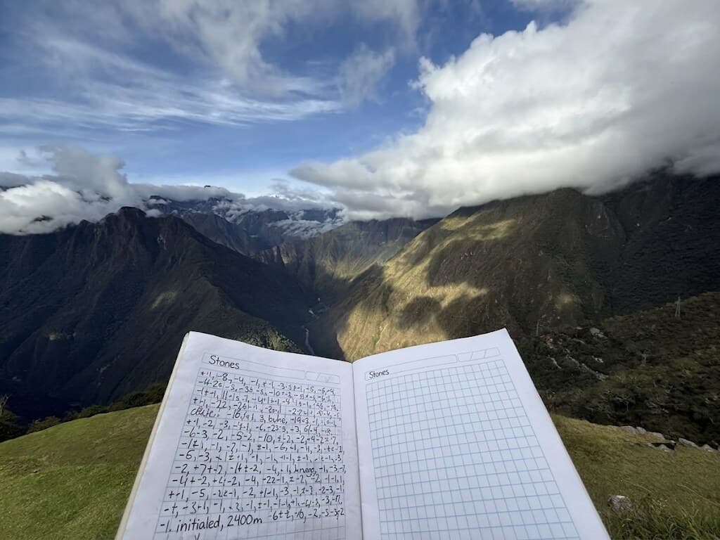 An open field notebook headed “Stones,” its grid pages filled with handwritten step tallies, propped on a grassy ridge with cloud-wrapped Andean peaks behind.